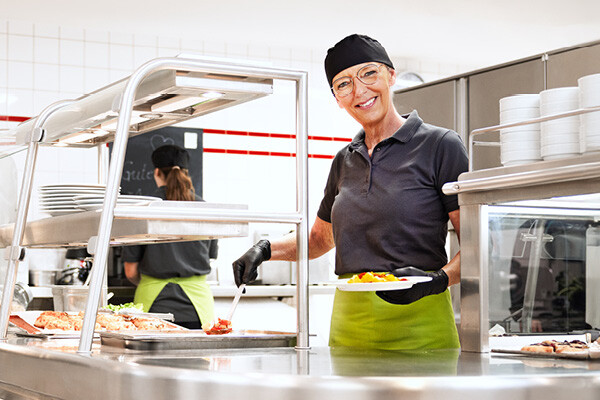 Start your career at Dussmann food services. Dussmann employee from the food service and hospitality division serving food in the cafeteria and smiling.