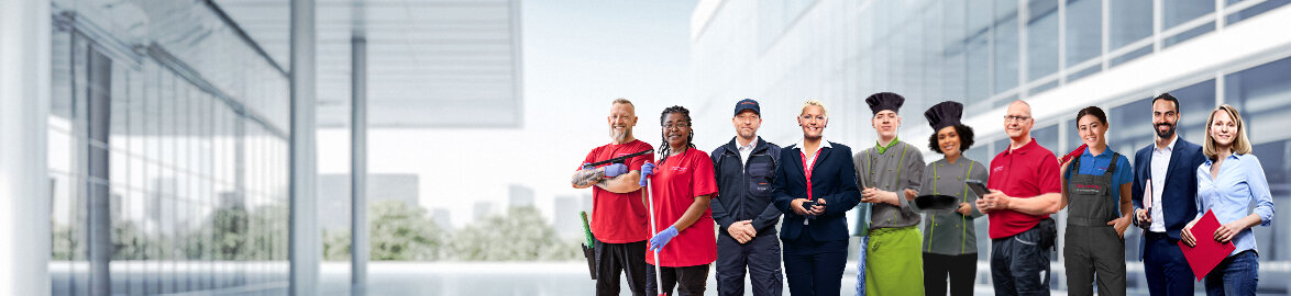 Dussmann bien plus qu'un employeur Photo de groupe d'employés souriants de Dussmann issus de différents domaines professionnels, devant un fond gris neutre