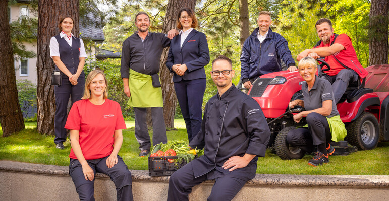 Become part of our Dussmann team – a wide variety of jobs await you. Group picture of smiling Dussmann employees from different work areas standing in a kind of large front yard with lawn and trees, wearing their work clothes.